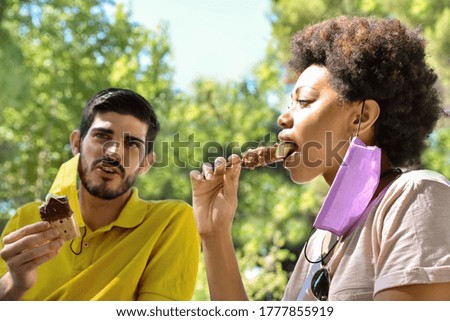 Interracial friends with a face mask hanging from their ear, who are enjoying ice cream, with a blurred trees background. Social gathering in pandemic times.