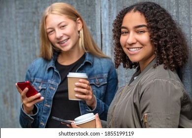 Interracial Friends Pretty Blonde Teenager And Mixed Race African American Girl Female Young Woman Using Their Smart Phones For Social Media And Drinking Takeout Coffee Outside