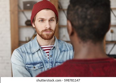 Interracial friendship concept. Handsome bearded European hipster wearing hat and denim jacket having serious conversation with his African American best friend, listening to him attentively - Powered by Shutterstock