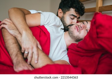 Interracial Gay Couple Passionate In Bed. Young Homosexual Partners In Red Bedding. LGBT Mixed Race And Caucasian Man Together Kissing In Bedroom Early Morning. 