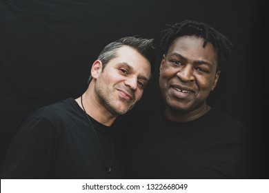 An Interracial Gay Couple Posing Before The Camera To Have Their Pictures Taken - Black Man And Mixed Race Man - On Black Background - With Copy Space. 