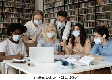 Interracial Group Of Gen Z Students Wearing Facial Medical Masks Sitting At Laptop In College Library, Watching Online Presentation, Discussing Learning Webinar, Working On Project Together