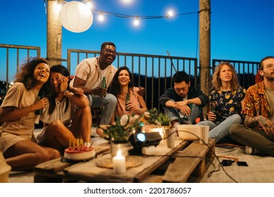 The Interracial Group Of People Sits On The Floor On The Rooftop And Watches The Football Game On The Projector. Some Of Them Are Happy About The Goal While Others Are Sad About Losing The Game.