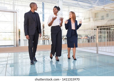 Interracial Group Of Professionals Chatting On Their Way To Lunch . Three Business People Walking Through Office Hall, Talking And Gesturing. Business Talks Concept