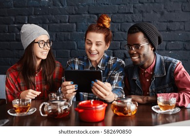 Interracial Group Of Three Hipsters Using Touch Pad At Cafe: Cute Caucasian Girl Holding Digital Tablet, Showing Something Funny On Internet To Asian Woman And African Man, Both In Hats And Glasses