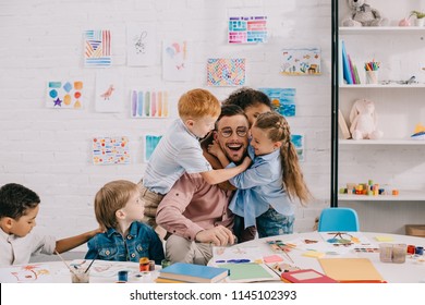 interracial kids hugging happy teacher at table in classroom