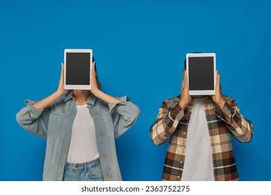 interracial man and woman obscuring face with digital tablets on blue backdrop, blank touch screen - Shutterstock ID 2363752275