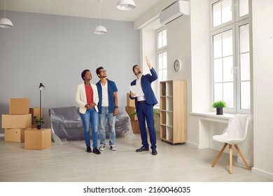 Interracial Married Couple Looking For New Home. Real Estate Agent Giving House Tour To Potential Buyers. Realtor Showing Modern Big Spacious Flat To Young Boyfriend And Girlfriend Or Husband And Wife