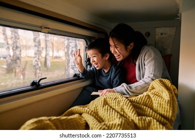 An Interracial Mother And Her Son Look Outside Through The Window Of Their Motor Home. They Lie On The Bed And Read The Book While The Child Is Waving.