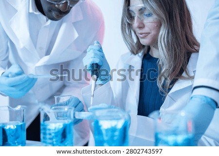 Interracial Scientist Duo Collaborates with Precision Pipette Over Beakers of Blue Solution - A Black male scientist and a blonde female colleague work in close collaboration around a lab bench, with the male scientist operating a precision pipette over a small beaker while the female leans in to observe. Vivid blue solution fills multiple beakers across the workspace.
