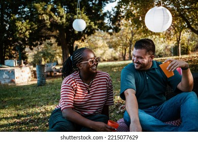 Interracial Smiling Couple Sitting On A Blanket In The Yard And Looking At Each Other. Multiracial Couple Having A Picnic On A Sunny Day In The Backyard