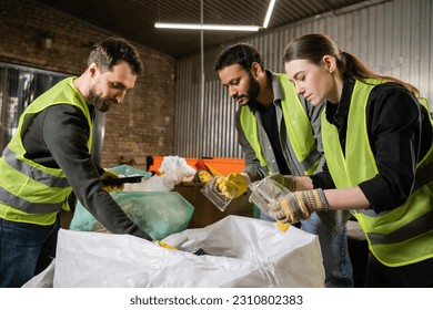 Interracial sorters in protective gloves and safety vests taking plastic containers from sacks while sorting trash together in waste disposal station, garbage sorting and recycling concept - Powered by Shutterstock