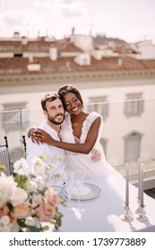 Interracial Wedding Couple. Destination Fine-art Wedding In Florence, Italy. African-American Bride And Caucasian Groom Are Sitting At The Rooftop Wedding Dinner Table Overlooking The City.