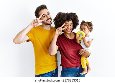 Interracial Young Family Of Black Mother And Hispanic Father With Daughter Doing Peace Symbol With Fingers Over Face, Smiling Cheerful Showing Victory 