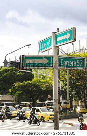 An intersection marked by clear signage becomes the center of a dynamic scene where the city flows without pause. Motorcycles, taxis, and pedestrians create an environment full of energy, while the signs visually guide the composition and convey order within the urban chaos.  
The green tones and natural light bring freshness, balancing the intensity of traffic with a sense of clarity and orientation.  
Ideal for mobility, transportation, tourism, urban planning projects, or content that seeks to reflect the real life of the city with authenticity and dynamism.