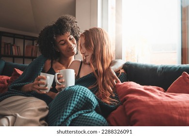 Intimate Moment for Lesbian Couple - An African American and a Caucasian woman with red hair embrace and cuddle on a sofa while drinking warm beverages from mugs, illuminated by a large window. - Powered by Shutterstock