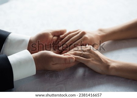 Intimate moment of two hands intertwined, showcasing a delicate engagement ring on a patterned tablecloth.
