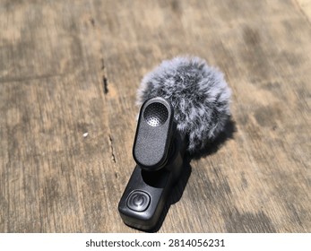An intimate, top-down view of a wireless lavalier microphone, highlighting its soft, grey faux-fur windscreen and the solid, matte black mounting clip. The device is placed on a rustic wooden surface, with natural textures adding depth to the composition. The image emphasizes the portability and ease-of-use of this modern audio tool, making it a perfect fit for visual assets related to vlogging, podcasting, and mobile content creation. The lighting is bright and directional, illuminating the intricate fur fibers and the clean lines of the microphone's chassis. This image is an excellent choice for commercial projects, technology blogs, e-commerce listings, or educational materials that require high-quality visuals of professional audio equipment. It effectively communicates the blend of modern design, functional performance, and reliability that is essential for modern multimedia producers.