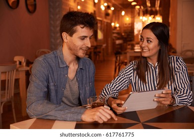 Intimately Smiling Millenial Couple Sitting At A Table In A Restaurant With Toothy Smiles While She Is Looking At Him Expectantly