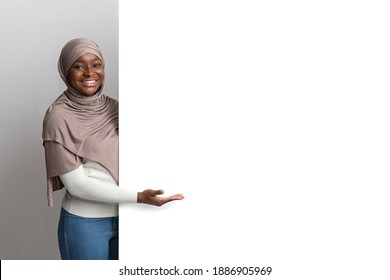 Intresting Offer. Happy Black Muslim Woman In Hijab Pointing At White Advertisement Board With Open Palm, Islamic Lady Showing Copy Space For Your Text Or Design, Standing Over Light Background