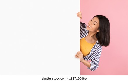 Intresting Offer. Happy Casual Asian Woman Peeping Out The Side Of White Advertisement Board And Holding It, Smiling Lady Looking At Copy Space For Your Text Or Design, Standing Over Pink Background