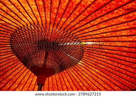 Intricate interior framework of an opened red oiled-paper umbrella (wagasa), showcasing the geometric symmetry of its woven bamboo ribs and vibrant, glowing red canopy illuminated by sunlight.