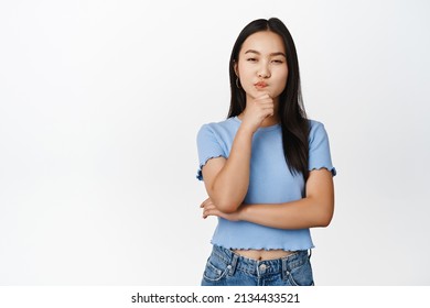 Intrigued asian woman looking thoughtful, thinking of something, having assumption, standing in blue tshirt over white background