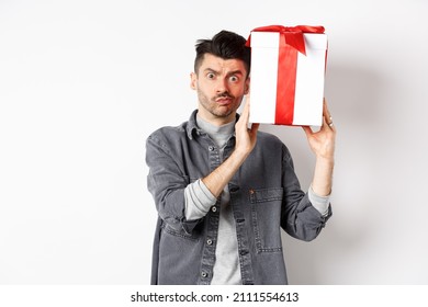 Intrigued Young Man Looking Confused At Camera, Trying To Guess What Inside Gift Box From Lover, Shaking Present And Stare Puzzled, Standing On White Background. Valentines Day Concept