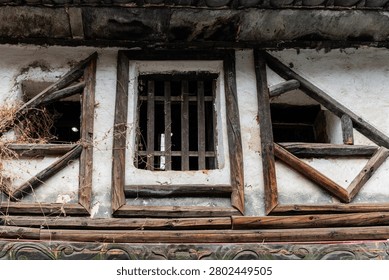 An intriguing architectural close-up of a rustic, weathered white plaster facade featuring three distinct traditional wooden window frames. The central opening is protected by sturdy vertical wooden bars, while the two flanking windows display unique diagonal geometric cross-bracing. Tangled dried vines cling to the left side of the aged timberwork, emphasizing the historic, timeworn character and authentic vernacular carpentry of rural southwestern China.