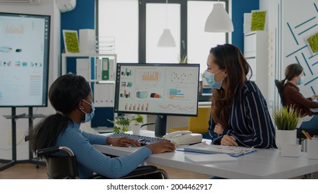 Invalid african businesswoman and employee with protective masks working together at financial project for start up company, compering statistics looking at computer discussing searching solution
