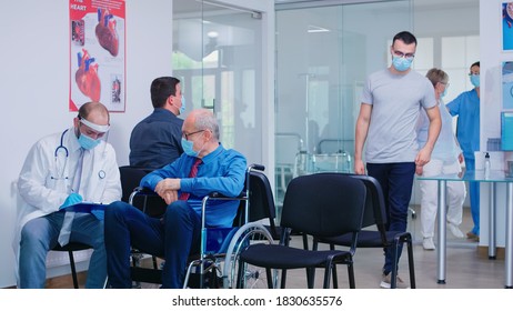 Invalid Old Man With Face Mask Against Infection With Coronavirus In Wheelchair Discussing With Doctor In Hospital Waiting Area. Nursing Inviting Patient In Examination Room.