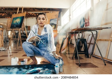 Inventive female painter looking at the camera in her art studio. Skilful artist making a blue painting for her new art project. Creative young woman working on the floor in her atelier.