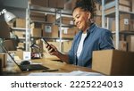 Inventory Manager Using Smartphone to Scan a Barcode on Parcel, Preparing a Small Cardboard Box for Postage. Black Multiethnic Small Business Owner Working on Laptop in Warehouse.