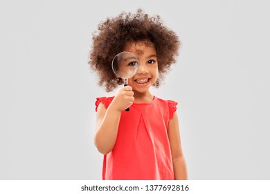 investigation, discovery and vision concept - happy little african american girl looking through magnifying glass over grey background - Powered by Shutterstock