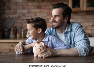 Investment in happy future. Young dad little tween boy sit at table hug hold piggy bank look at distance dream of wealth. Adult single dad embrace preteen son plan trip big purchase after saving money