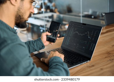 Investor using mobile phone and laptop checking trade market data. Stock trader broker looking at computer analyzing trading cryptocurrency finance market crypto stockmarket data, over shoulder view. - Shutterstock ID 2339480179