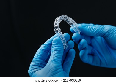 Invisible orthodontics cosmetic aligners in hands of doctor wearing sterile gloves isolated on a black background, tooth aligners, plastic braces. Modern teeth retainers created on a 3d printer.