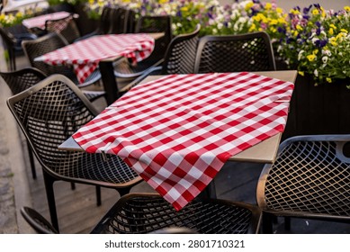 An inviting empty restaurant patio setup featuring traditional red-checked table runners clipped onto tables next to vibrant summer flowers