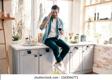 Inviting Friends For Dinner. Handsome Young Man In Casual Wear Drinking Wine And Talking On The Phone While Sitting In The Kitchen At Home