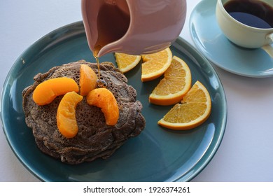 Inviting Stack Of Healthy Buckwheat Pancakes With Grilled Peach And Fresh Orange Slices, Coffee, And Pitcher Above Plate Pouring Maple Syrup. 