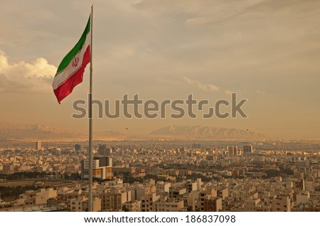 Iran flag waving  in the wind above skyline of Tehran lit by orange glow of sunset.