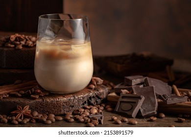 Irish Cream And Coffee Cocktail In A Glass With Ice On An Old Wooden Background. Coffee Beans, Cinnamon, Anise, And Pieces Of Chocolate Are Scattered On The Table. 