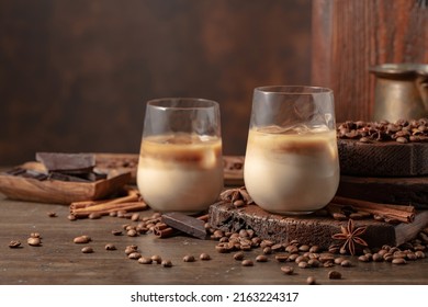Irish cream and coffee cocktail in glasses with ice on an old wooden background. Coffee beans, cinnamon, anise, and pieces of chocolate are scattered on the table. 