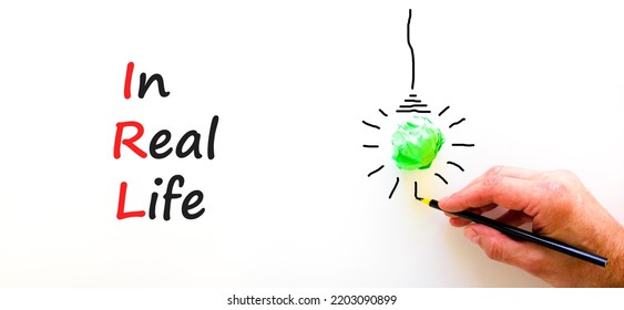 IRL In Real Life Symbol. Concept Words IRL In Real Life On White Paper On A Beautiful White Background. Green Light Bulb Icon. Businessman Hand. Business And IRL In Real Life Concept. Copy Space.