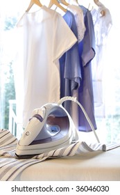 Iron On Ironing Board With Clothes Hanging In Background