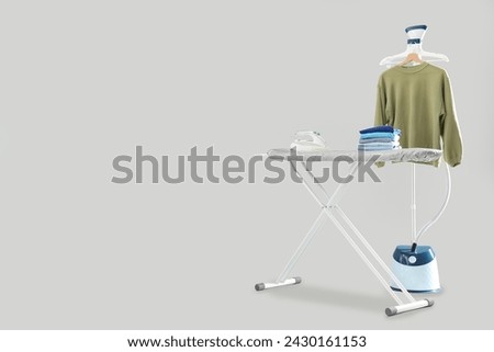 Ironing board with stack of clean clothes and garment steamer on grey background