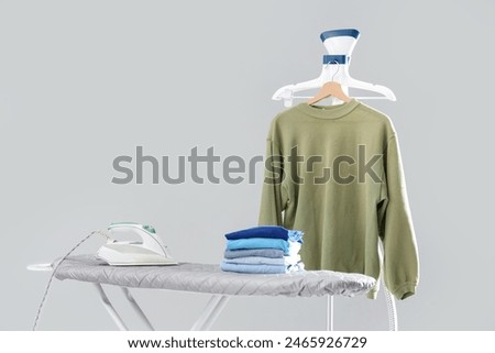 Ironing board with stack of clean clothes and garment steamer on grey background