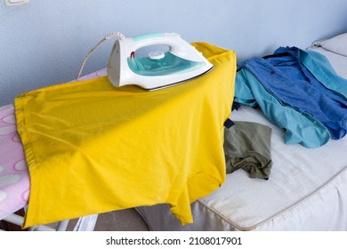 Ironing clothes on a yellow T-shirt on the ironing board and in the background more clothes to be ironed on the bed. Concept of housework.
