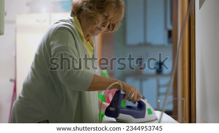 Ironing Out Domestic Chores_ Candid Footage of Senior Woman in Laundry Room. Authentic real life older lady doing everyday routine, ironing clothes with steam iron