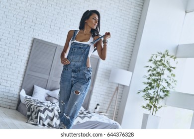 Irresistible Girl. Attractive Young Woman Touching Her Bib Overalls And Looking Away While Walking In The Bedroom At Home